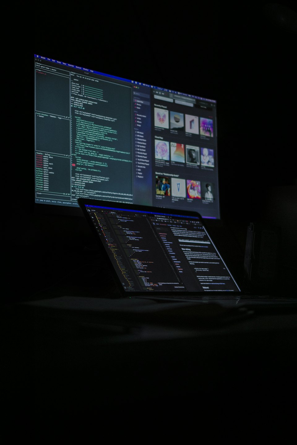 Laptop and monitor displaying complex code in a dark room.